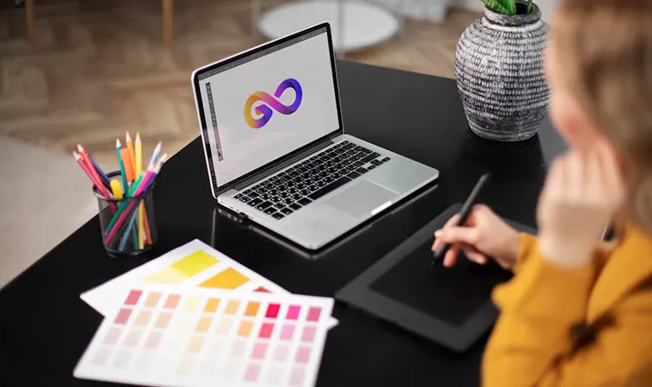 Person working at a desk with a laptop displaying an infinity symbol, color swatches, pencils, and a tablet.